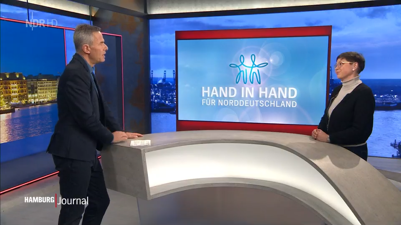 Fernsehstudio Hamburg Journal: Gesa Müller und Moderator stehen sich gegenüber, im Hintergrund ein Bildschirm auf dem das Logo der Aktion "Hand in Hand für Norddeutschland" ist