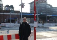 Ein Mann steht an einer Bushaltestelle, man sieht den Zaun einer Baustelle