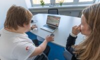 Eine Person sitzt am Laptop. Eine andere Person sitzt neben ihr und schaut zu.