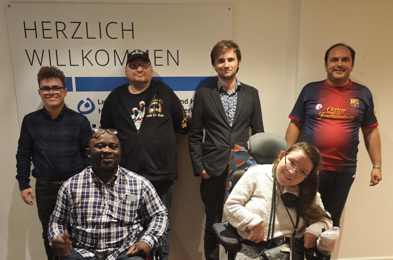 Gruppenfoto der 6 Peer-Berater: 5 Männer und eine Frau stehen zusammen und lächeln freundlich. 2 Personen haben einen Rollstuhl.